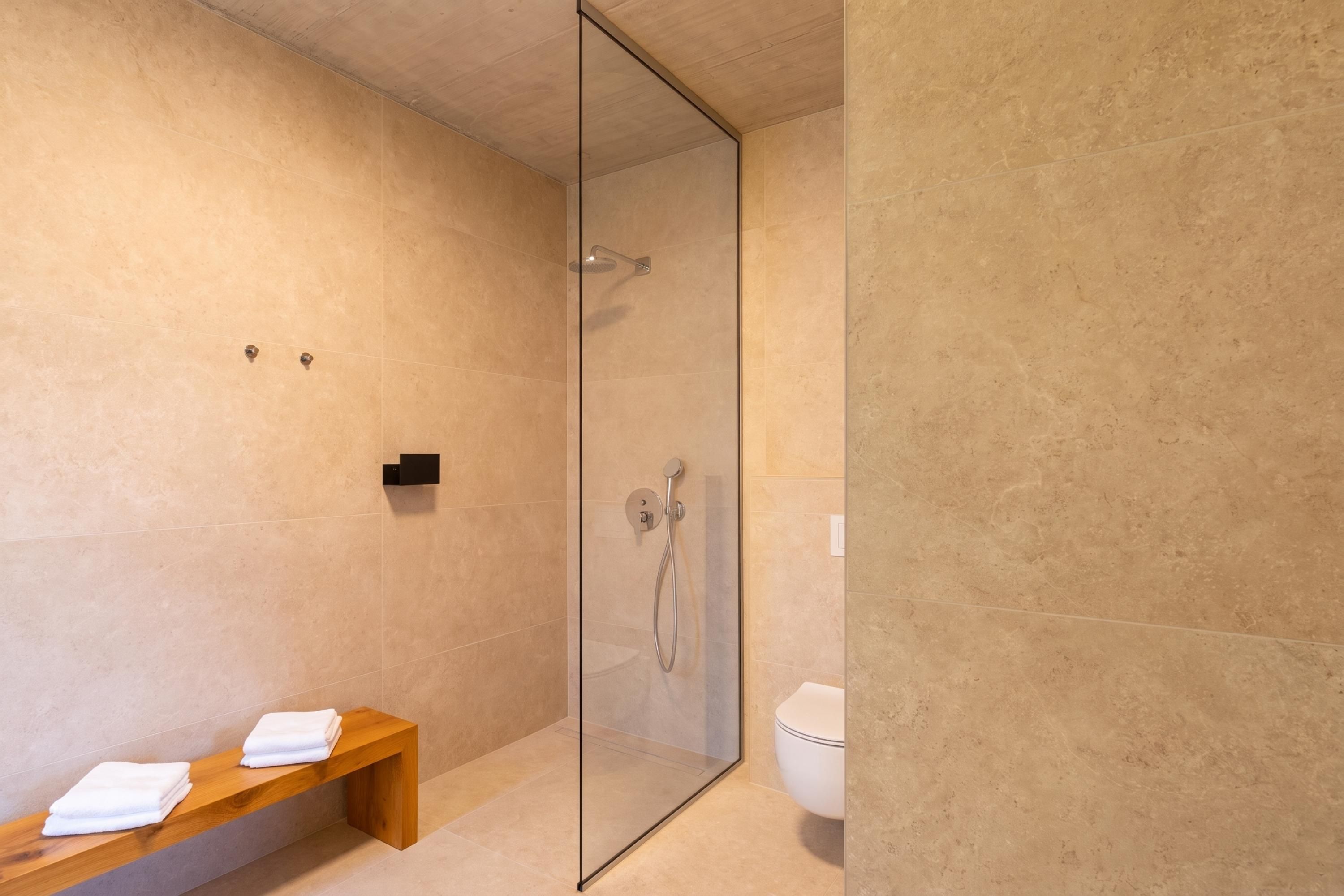 A modern bathroom with a glass shower enclosure, a wooden bench with towels, and a toilet.