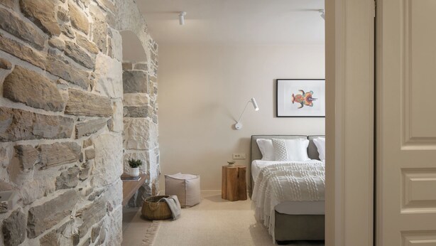 A bedroom with a stone wall, a bed with white bedding, a bedside table with a lamp, and a framed picture on the wall.