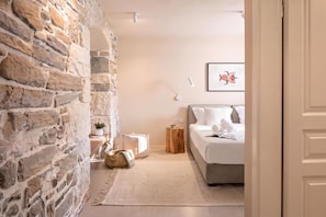 Un dormitorio con pared de piedra, una cama con ropa de cama blanca, una mesita de noche pequeña, una lámpara y un cuadro enmarcado en la pared.