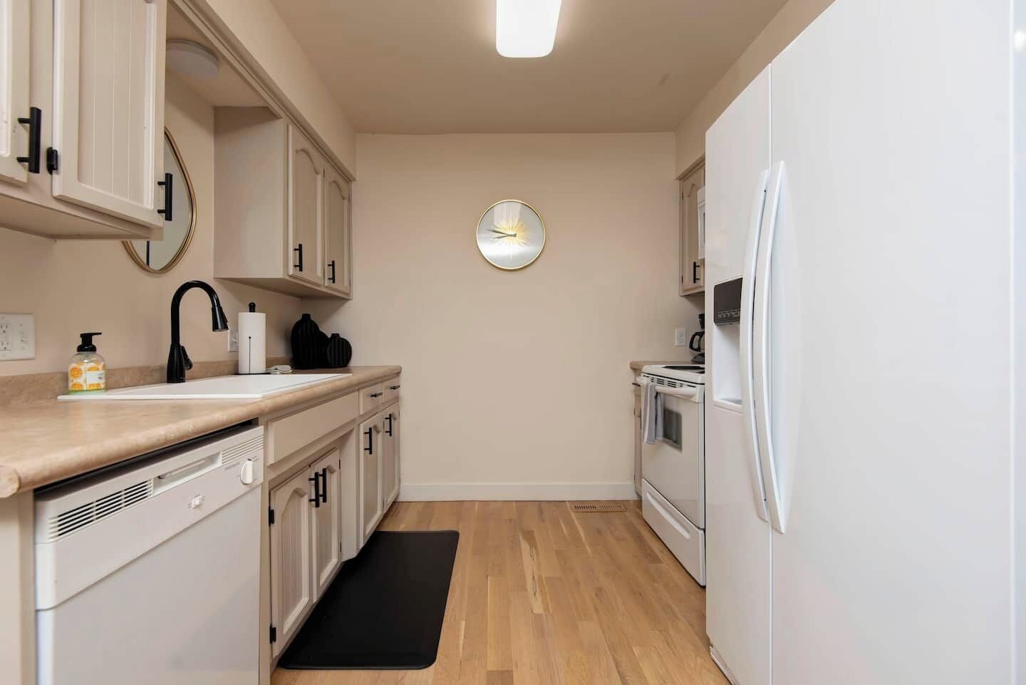 Una cocina compacta con armarios blancos, un grifo negro, un reloj en la pared y un refrigerador blanco.