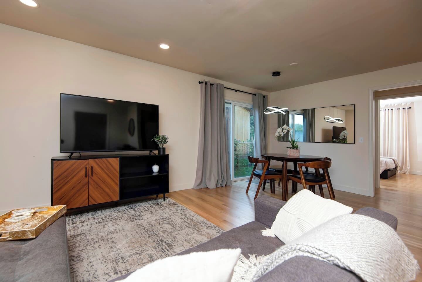 A modern living room with a flat-screen TV, wooden entertainment unit, grey sofa, dining table with chairs, and a large mirror.