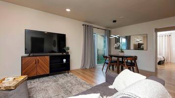 A modern living room with a flat-screen TV, wooden entertainment unit, grey sofa, dining table with chairs, and a large mirror.