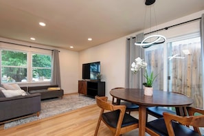 A modern living room with a sofa, TV, dining table, chairs, and a large window with curtains.