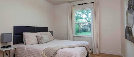 Un dormitorio con cama, mesita de noche, lámpara, ventana con cortinas y un cuadro en la pared.