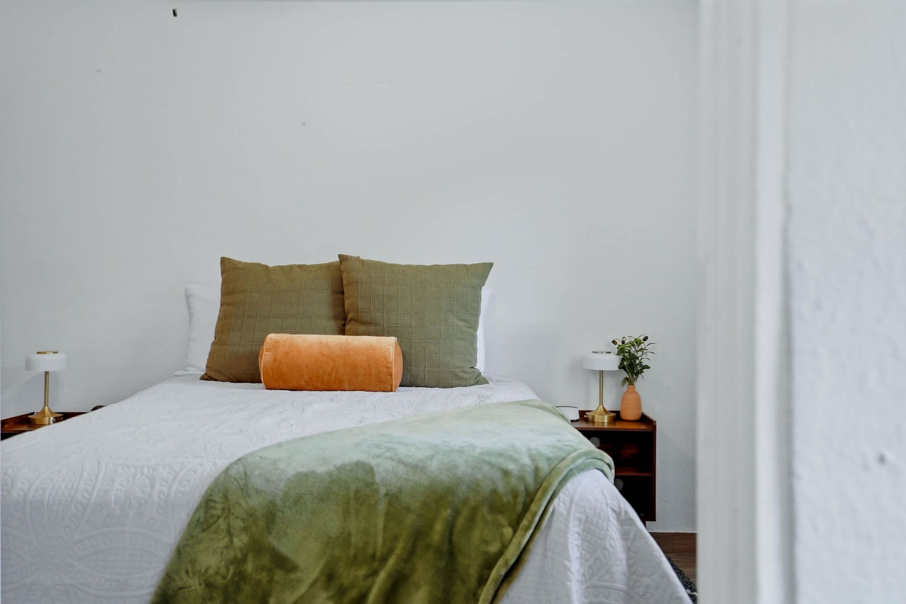Una cama bien hecha con una manta verde, dos almohadas y una lámpara de noche sobre una mesita de madera.