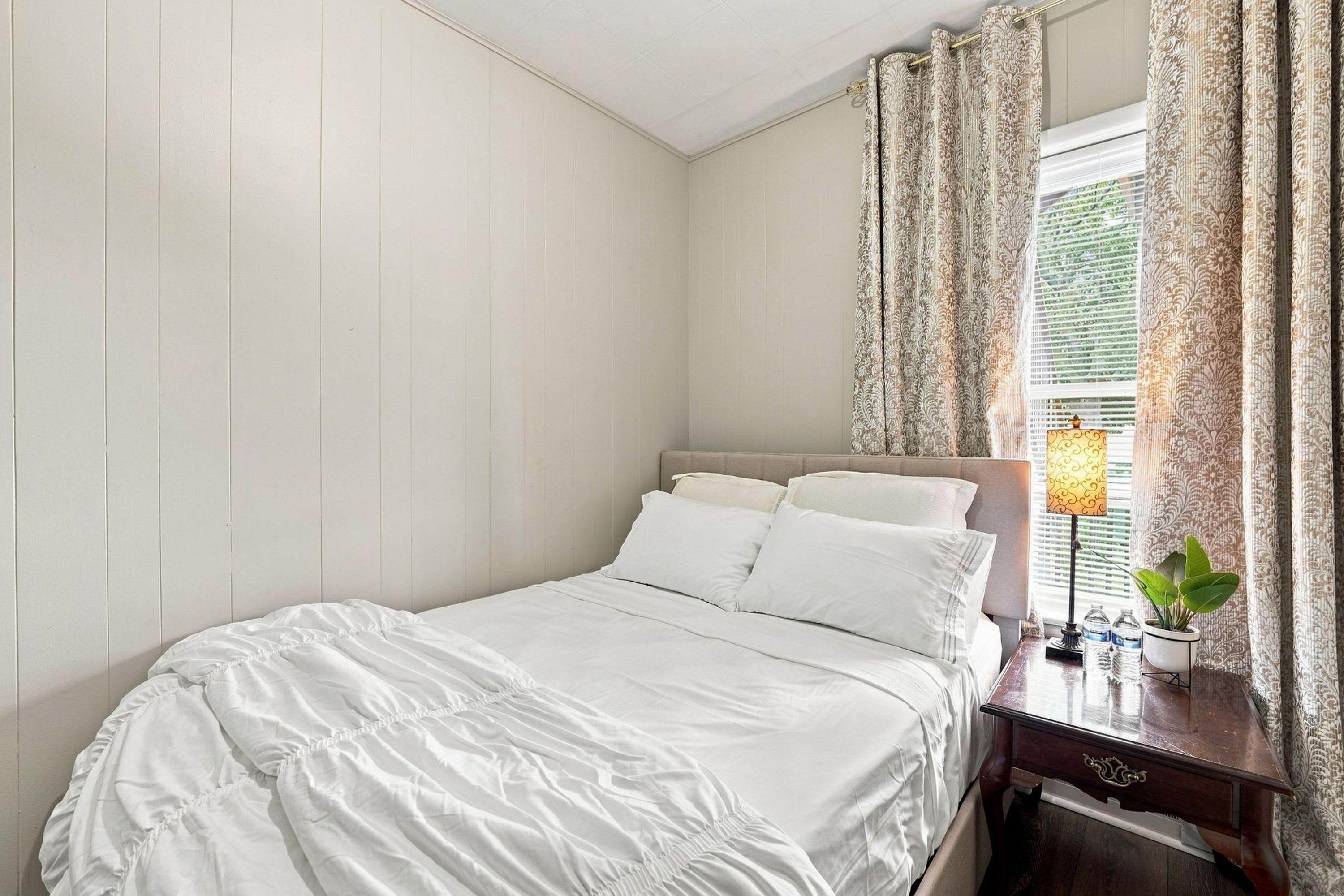 Una cama bien hecha con sábanas blancas en una habitación de paredes claras y una ventana con cortinas estampadas.