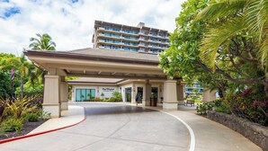 Entrada de un hotel con un corredor cubierto, palmeras y un edificio al fondo.
