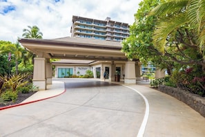 A hotel entrance with a covered walkway, palm trees, and a building in the background. - Whaler | Ocean View Studio Sleeps 2 | Car Incl. w/ 6+ Nights | WH2-465 by KBM (Lahaina)