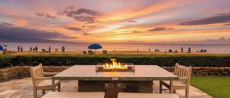 A patio with a fire feature, seating, and a view of the beach at sunset.