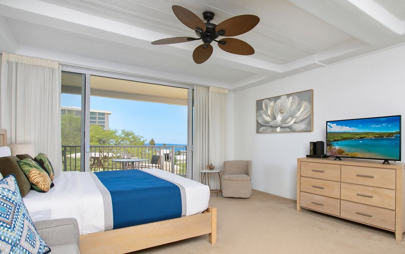 Hotelkamer met een bed, televisie, plafondventilator, balkon met uitzicht en een klein tafeltje met een stoel.