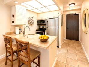 A kitchen with a central island, wooden chairs, a microwave, oven, refrigerator, and a door with a circular mirror. - Whaler | Ocean View Studio Sleeps 2 | Car Incl. w/ 6+ Nights | WH2-465 by KBM (Lahaina)