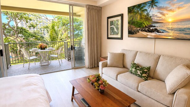 Habitación de hotel con balcón, sofá, cama, una mesita de centro con flores y un cuadro de una playa.