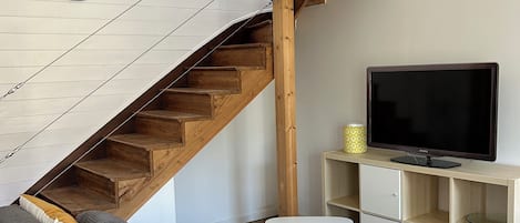 Una sala de estar con una escalera de madera, un sofá gris, un mueble blanco para televisor y una pequeña mesa blanca.
