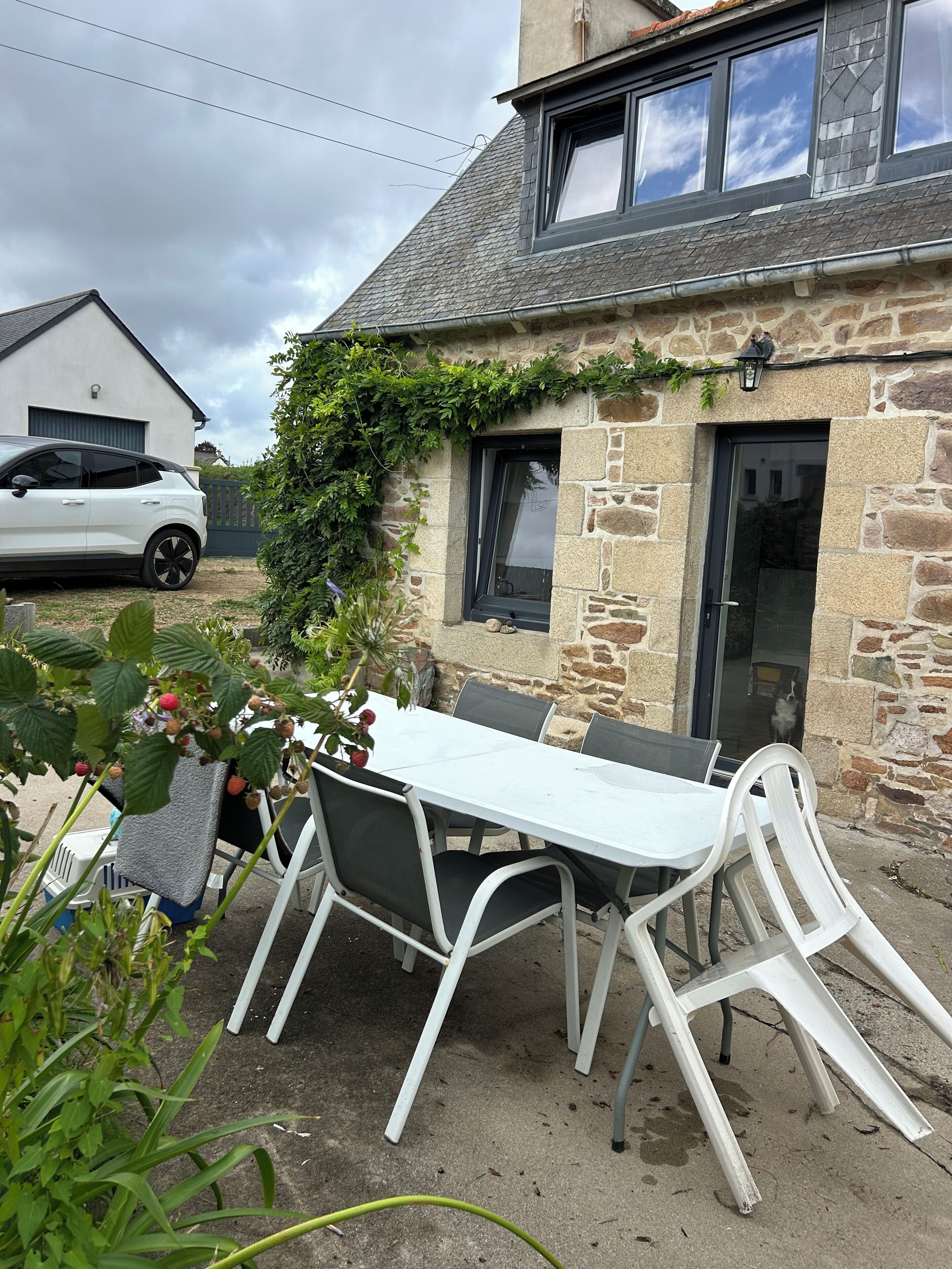 A stone building with a white table and chairs outside, a car parked nearby, and a dog visible through the door.