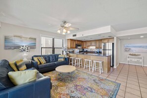 A modern living room with a blue sofa, a patterned rug, and a kitchen area with wooden cabinets and a stainless steel refrigerator. - Phoenix II 2031 by Liquid Life (Orange Beach)
