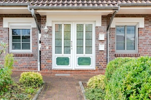 A two-story house with a brick facade, a white front door with glass panels, and a small garden area with shrubs.