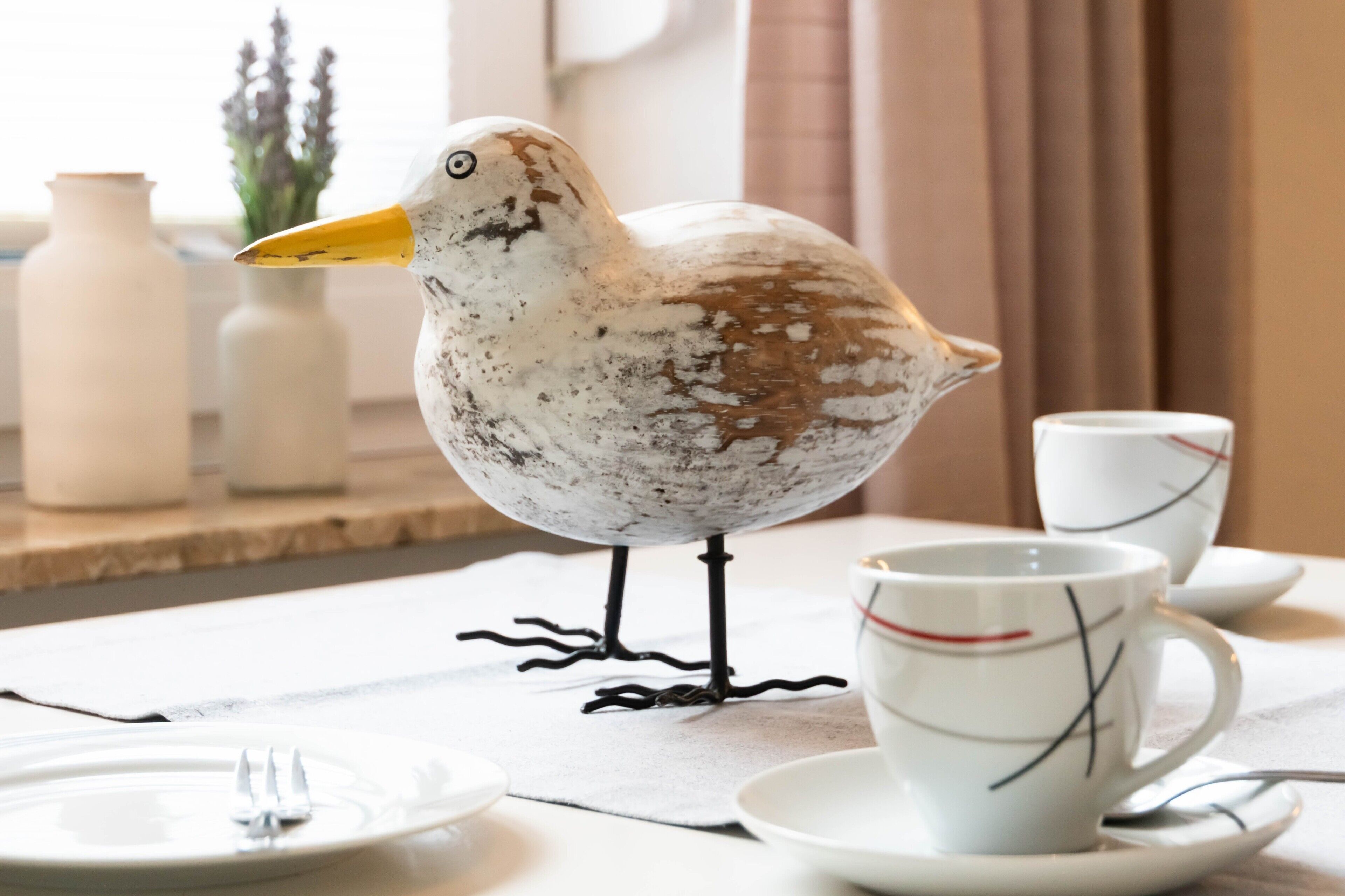 Una figurita de pájaro sobre una mesa con una taza y un platillo, un tenedor, y un florero al fondo.