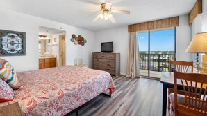 Habitación de hotel con cama, escritorio, silla, televisor, ventilador de techo y balcón con vistas.