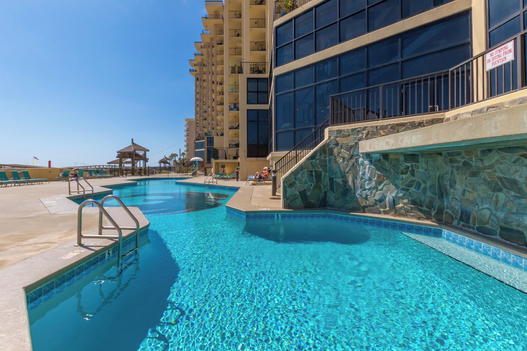A swimming pool with clear blue water, surrounded by lounge chairs and a stone wall.