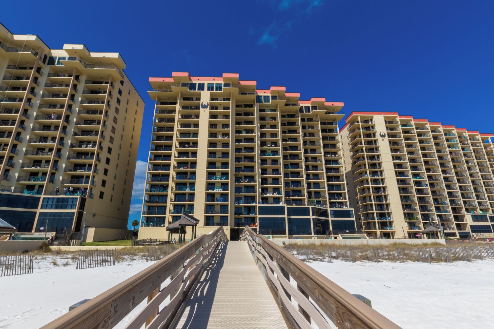 A boardwalk leading to high-rise buildings with balconies, a sandy beach, and a clear blue sky.
