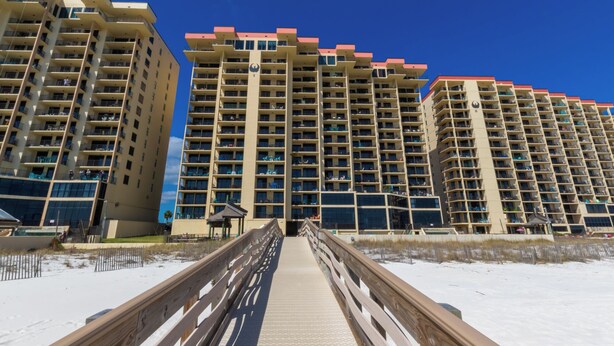 Un paseo marítimo que conduce a edificios altos con balcones, una playa arenosa y un cielo azul despejado.