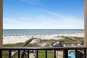 Blick auf einen Strand vom Balkon mit einer Holzdielen-Terrasse, einem Pool und Liegestühlen.