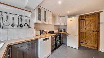 A compact kitchen with white cabinets, a dishwasher, a stove, and a refrigerator. There is a wooden door and a hanging utensil rack.