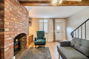 A cozy living room with a brick fireplace, a green armchair, a grey sofa, and a wooden floor. - Quaint Cottage in Charming Evesham (Evesham)