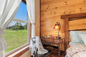 Ein Zimmer mit Holzwand, einem Fenster mit weißen Vorhängen, einem Schreibtisch mit Lampe und einem Bett mit gemustertem Überwurf.