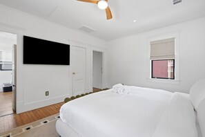 A hotel room with a large bed, a flat-screen TV mounted on the wall, and a window with a blind. - The Magnolia Suite at One 01 (Savannah)