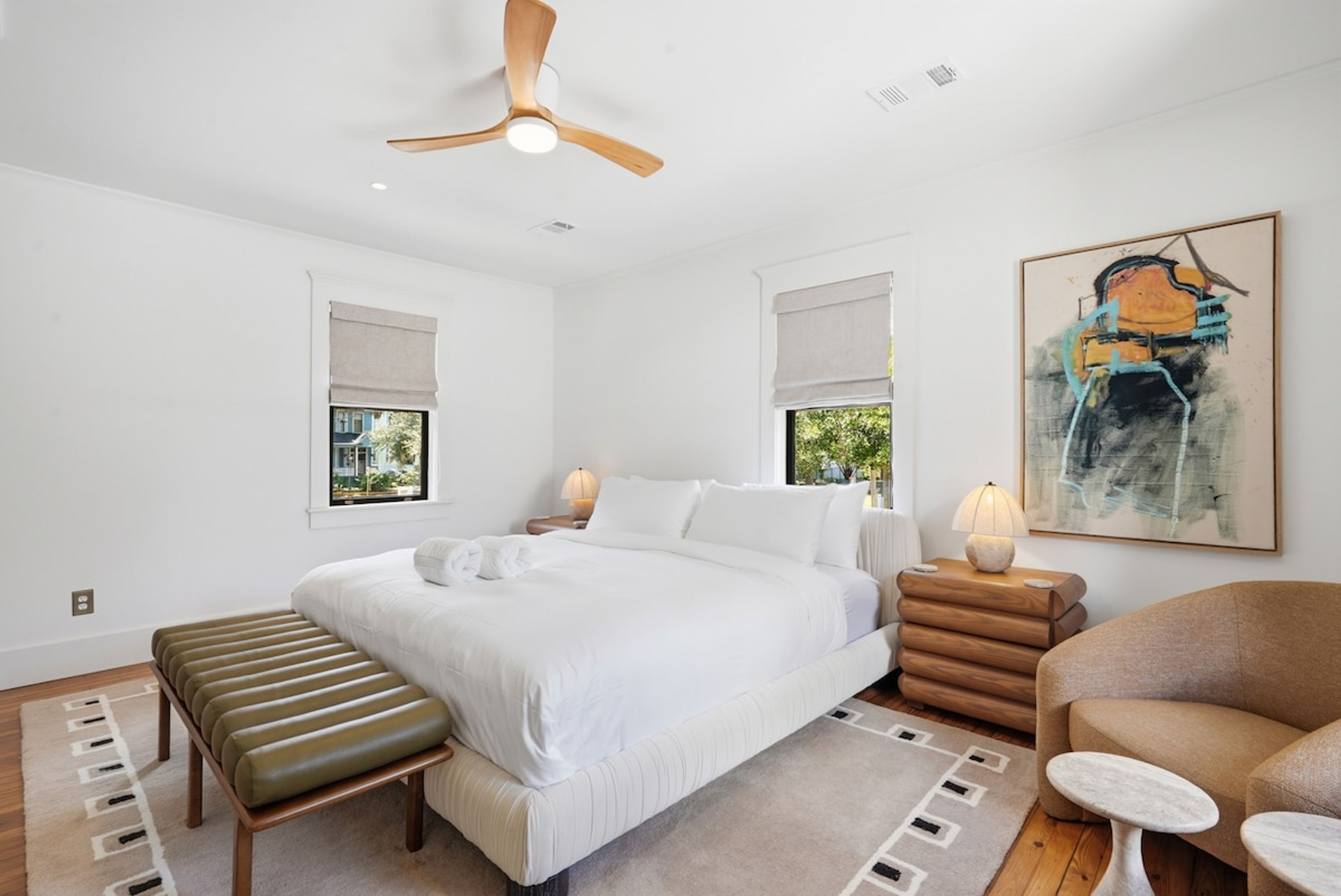 A bedroom with a bed, bedside tables, a bench, a chair, a ceiling fan, and a painting.
