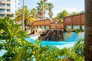 A pool area with a wooden bridge, palm trees, and a building in the background. - Paradise at The Palms | Gulf Views, Resort Pool & Beach Gear (Destin)