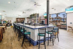 A modern bar area with a central island, bar stools, and a view of the beach through large windows. - Paradise at The Palms | Gulf Views, Resort Pool & Beach Gear (Destin)