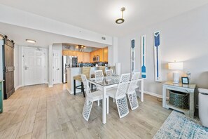 A modern kitchen with wooden cabinets, stainless steel appliances, and a dining area with white chairs and a glass table. - Paradise at The Palms | Gulf Views, Resort Pool & Beach Gear (Destin)