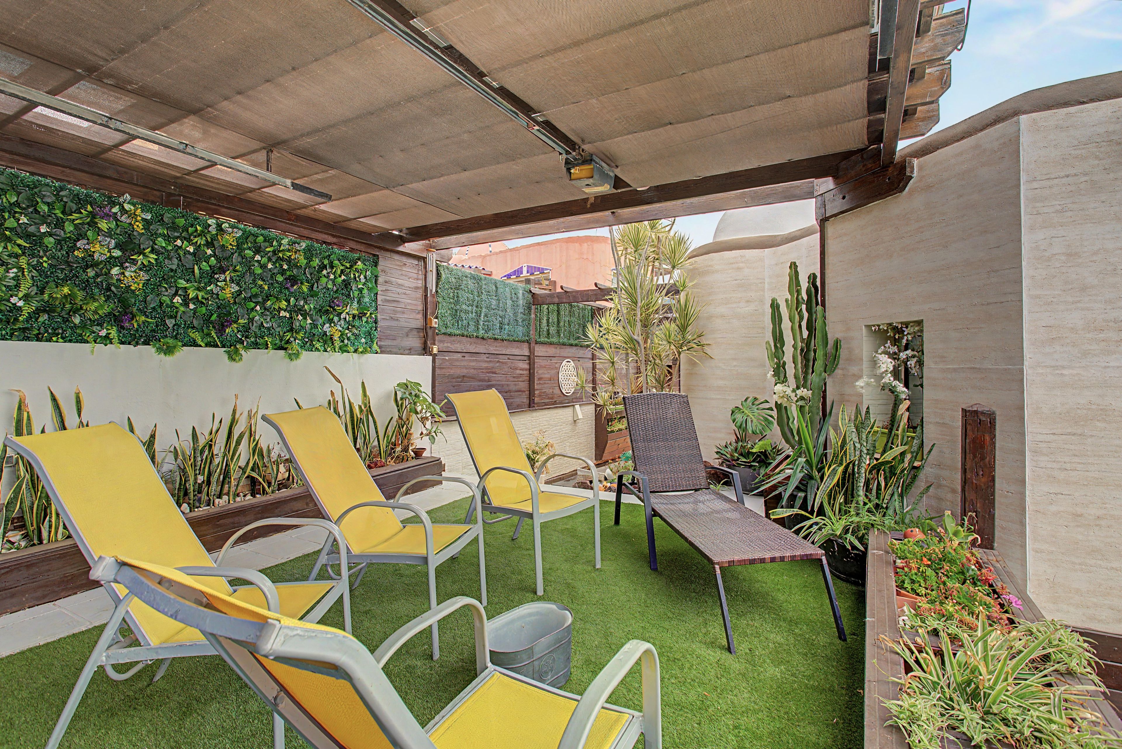 Un espacio al aire libre con patio, césped artificial, sillones reclinables amarillos y grises, y varias plantas en macetas.