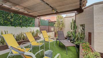 Un espacio al aire libre con patio, césped artificial, sillones reclinables amarillos y grises, y varias plantas en macetas.