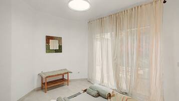 Una habitación con un banco de madera, una cama con toallas enrolladas, una ventana con cortinas traslúcidas y una lámpara en el techo.