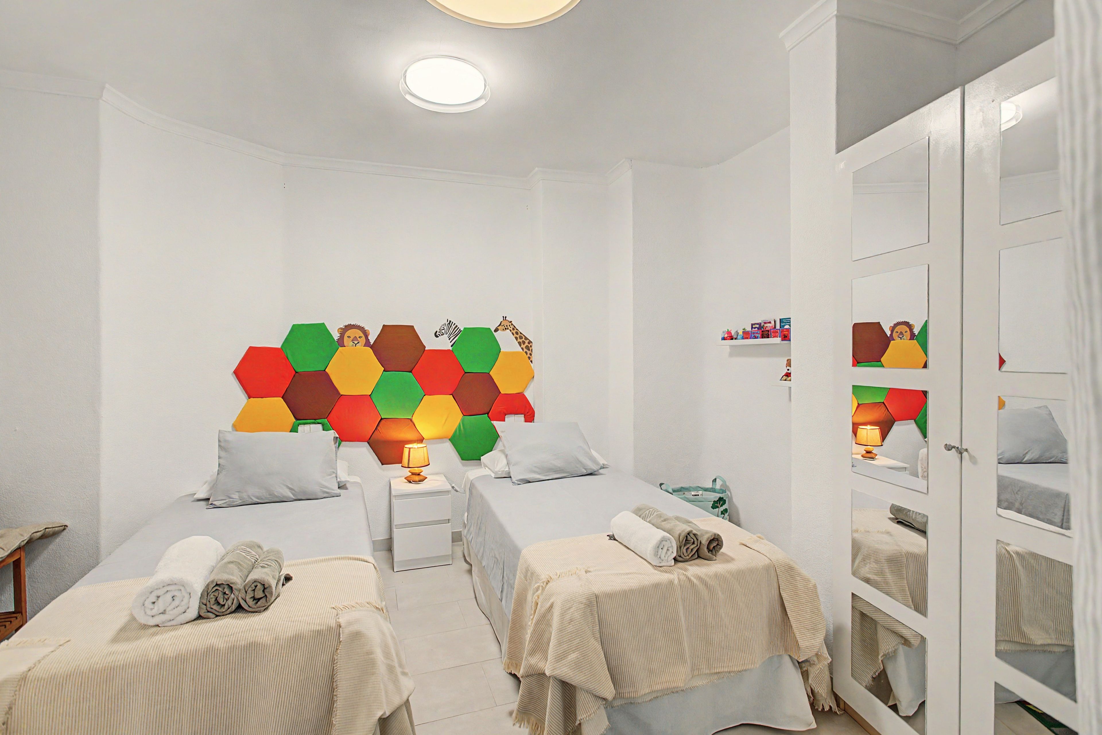 A hotel room with two beds, a colorful hexagonal wall decoration, a small table with a lamp, and a wardrobe with mirrors.
