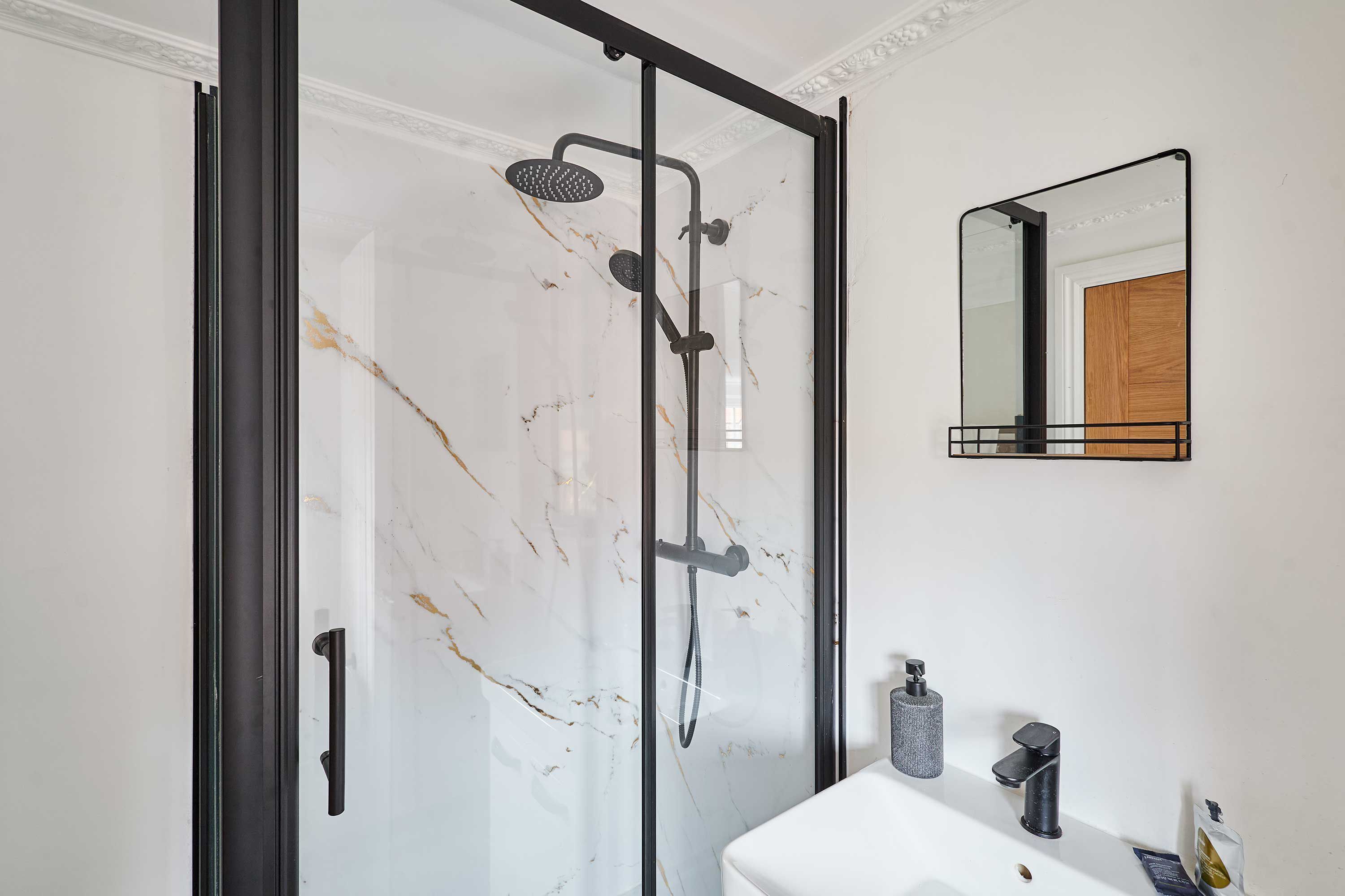 Une salle de bain moderne avec une douche à parois de verre, un mur en marbre blanc, une porte en bois, un miroir à cadre noir et un lavabo blanc doté d’un distributeur de savon.