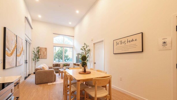 A modern, well-lit living room with a dining area, large windows, and a view of the outdoors.