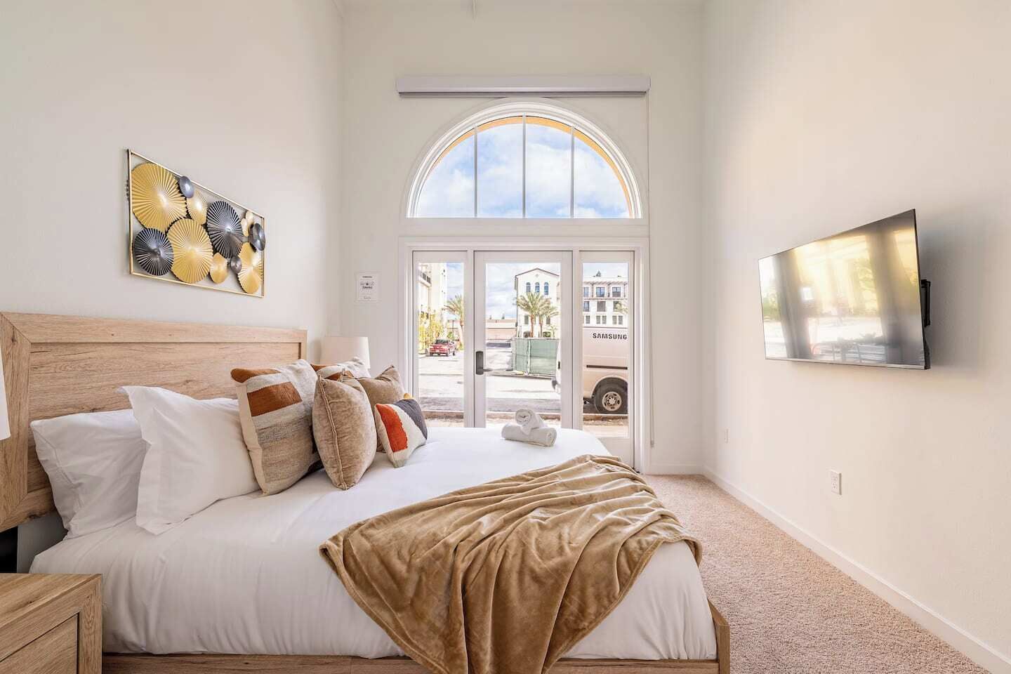 A bedroom with a bed, pillows, a blanket, a TV, and a window with a view of the outside.