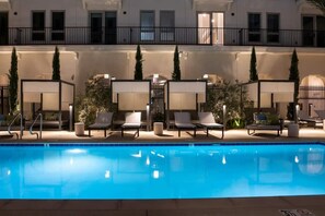 Una piscina con sillas de descanso y sombrillas, rodeada por un edificio con balcones y una valla.