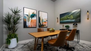 A modern dining area with a wooden table, four chairs, abstract paintings, a large TV, and a potted plant.