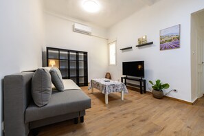 Ein modernes Wohnzimmer mit einem grauen Sofa, einem kleinen Tisch mit Lampe, einem Fernseher auf einem Standfuß und einer Topfpflanze.