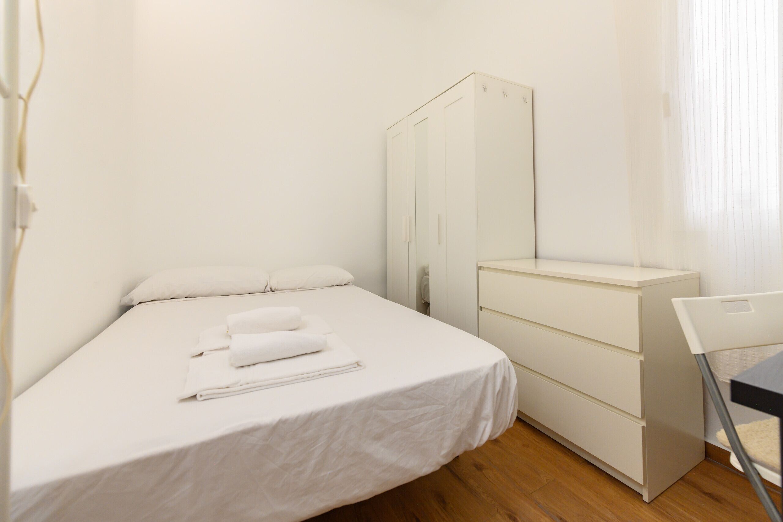 Quarto individual com uma cama branca, uma pequena escrivaninha e um guarda-roupa.