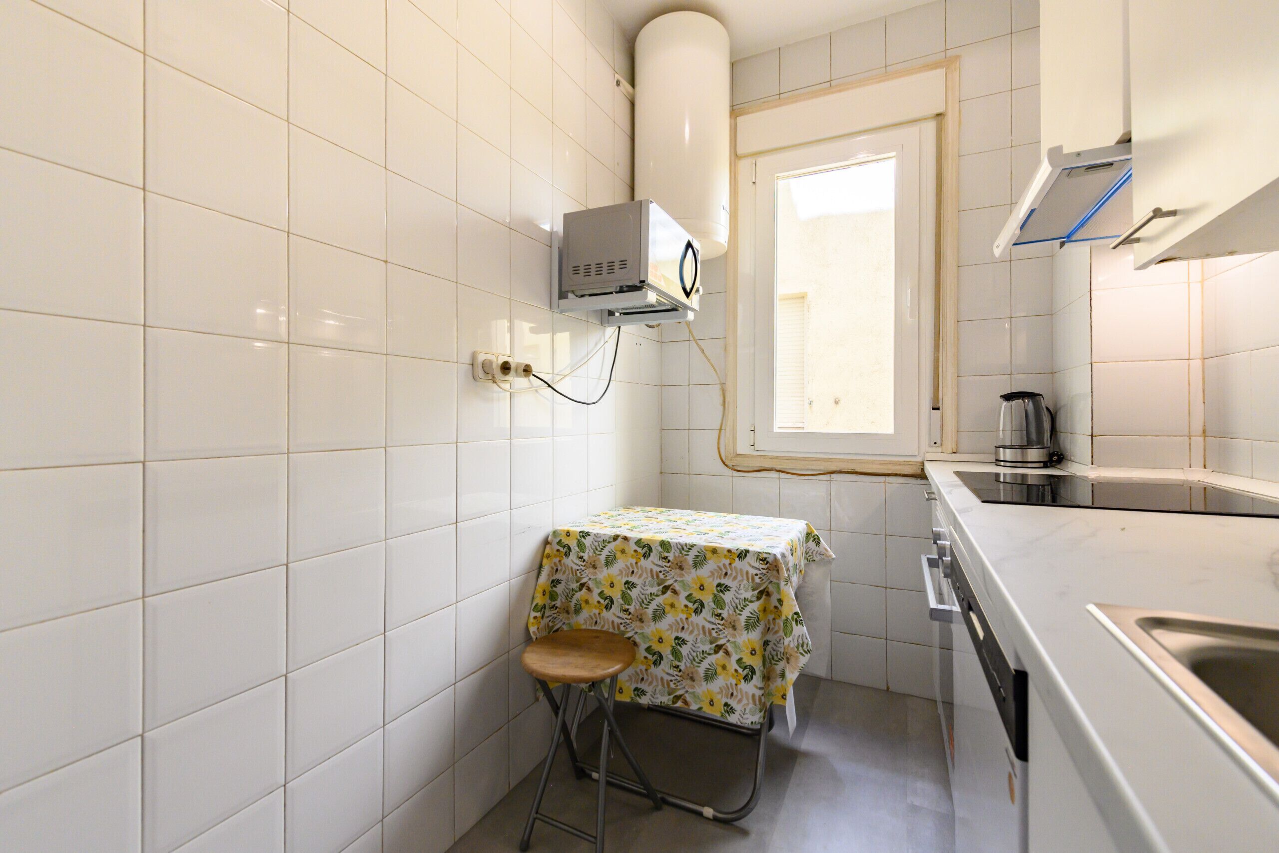A small kitchen with white tiled walls, a window, a table with a floral tablecloth, and a sink.