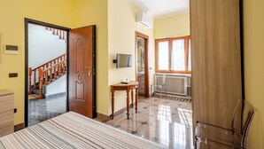 A hotel room with a bed, a wooden door, a small table, a chair, a TV, and a window with curtains.