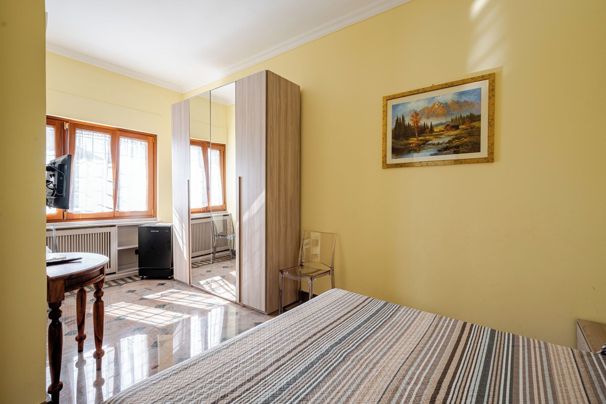 A bedroom with a bed, a chair, a desk with a computer, a radiator, and a painting on the wall.