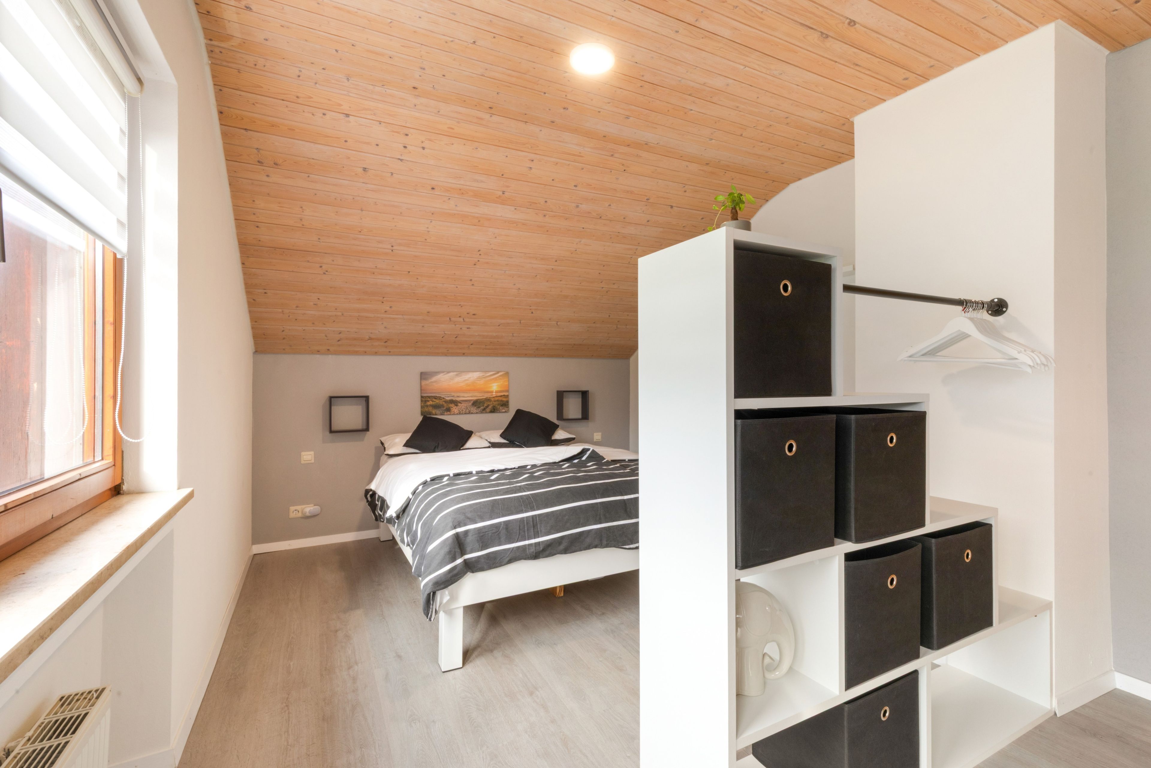 Ein Schlafzimmer mit einem Bett, einem Fenster, einer Decke mit Holzbalken und einer an der Wand befestigten Ablage.