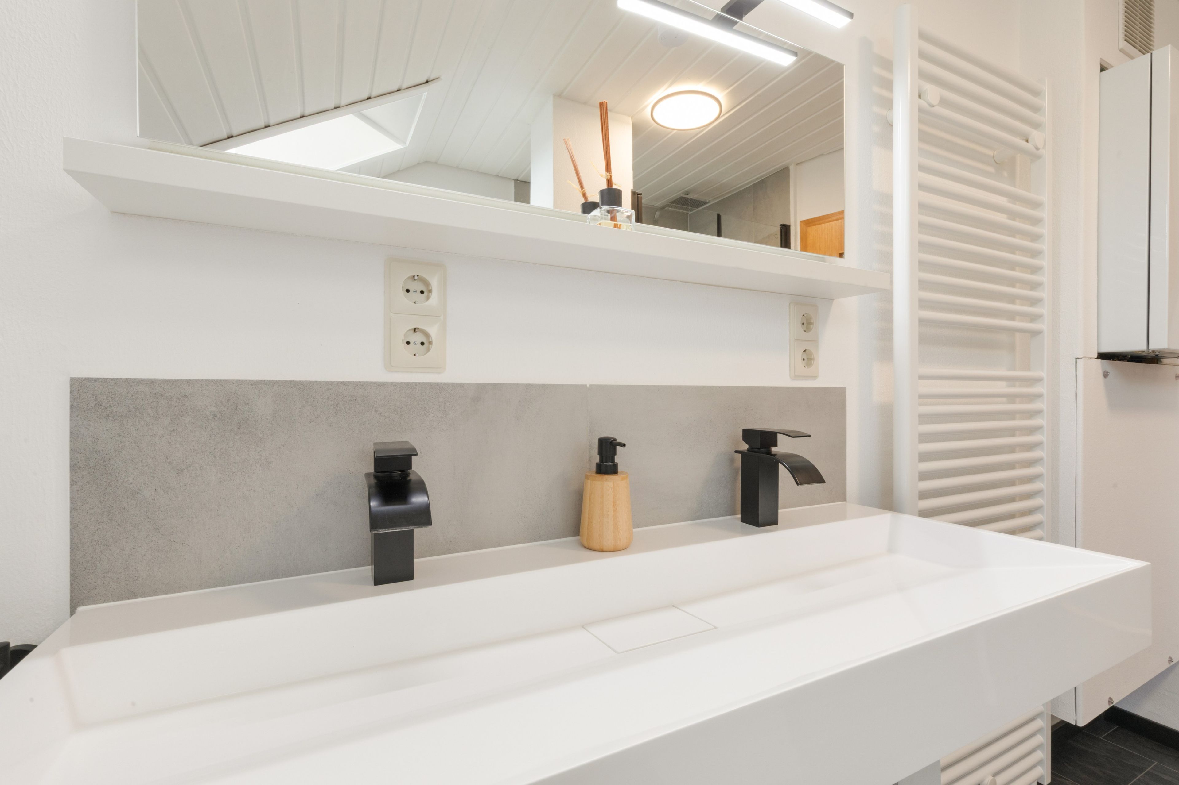 Une salle de bain moderne avec un meuble-lavabo blanc, deux robinets noirs, un distributeur de savon en bois et un miroir au-dessus du lavabo.